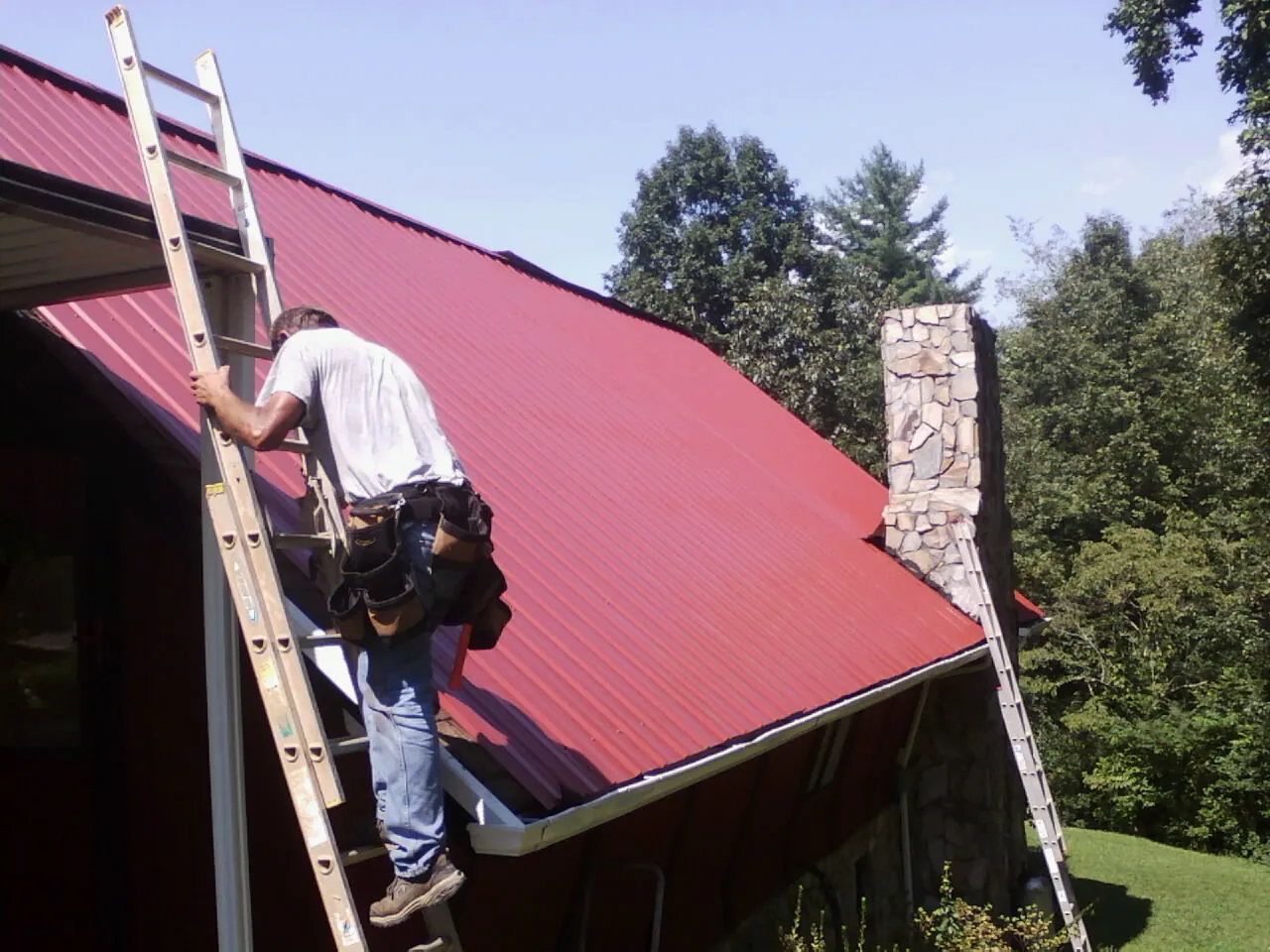 Roofer on a ladder beside a red metal roof and stone chimney — Soffit & Fascia Repair by Roofing Brothers in Divide